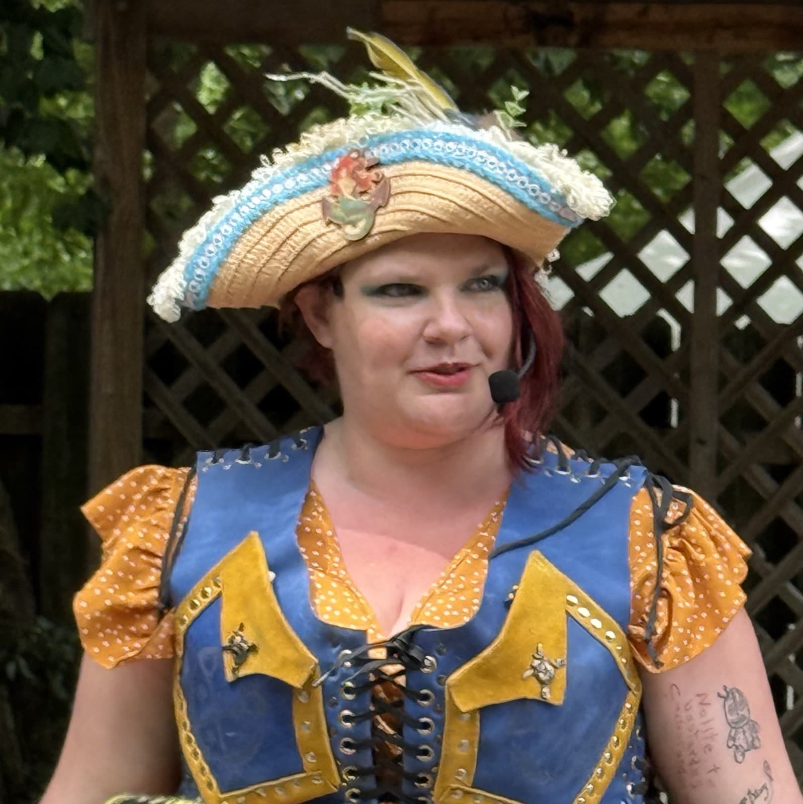 Erica Minton as Captain Shh-nel: a pirate captain in a wide feather-trimmed tricorn hat and blue-and-gold laced bodice, wearing a headset mic, in front of a lattice fence