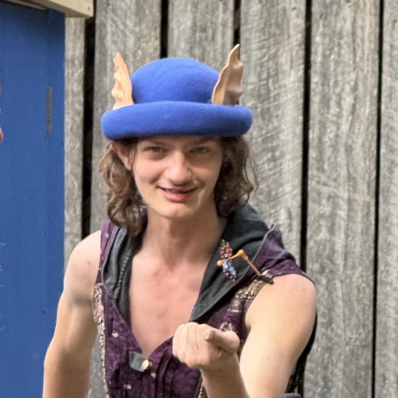 Phoenix Minton as Stu of the Loo: a sewer-rat character in a blue felt hat with tan rat ears and a purple vest, raising a fist toward the camera, in front of weathered wood planks
