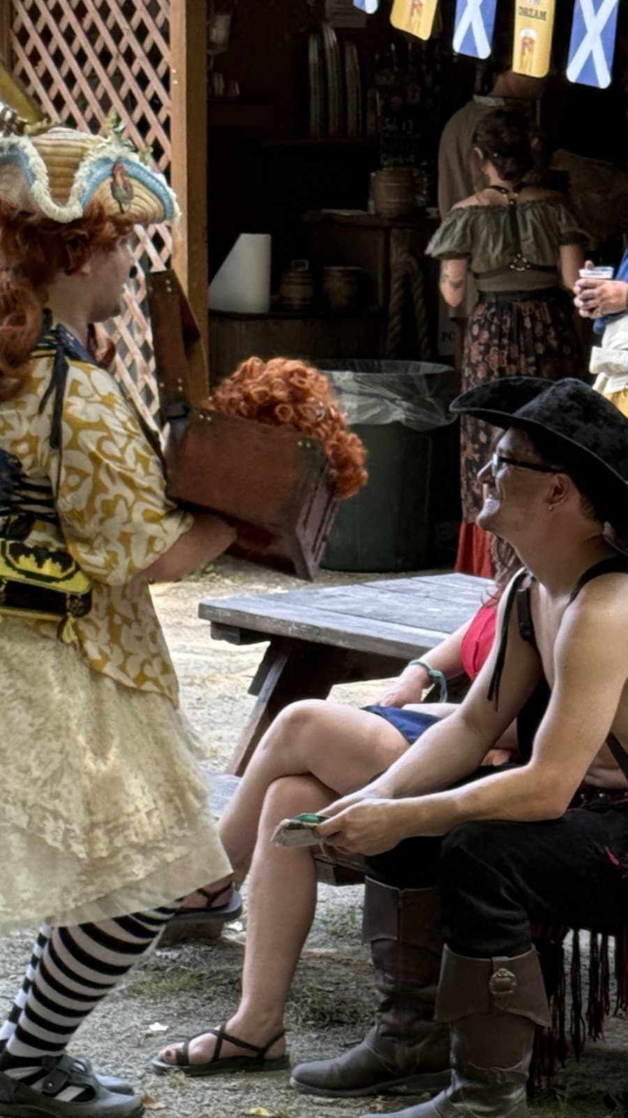 A costumed Shh It's A Show performer serenades a seated patron at an outdoor faire stall, a wooden box in hand