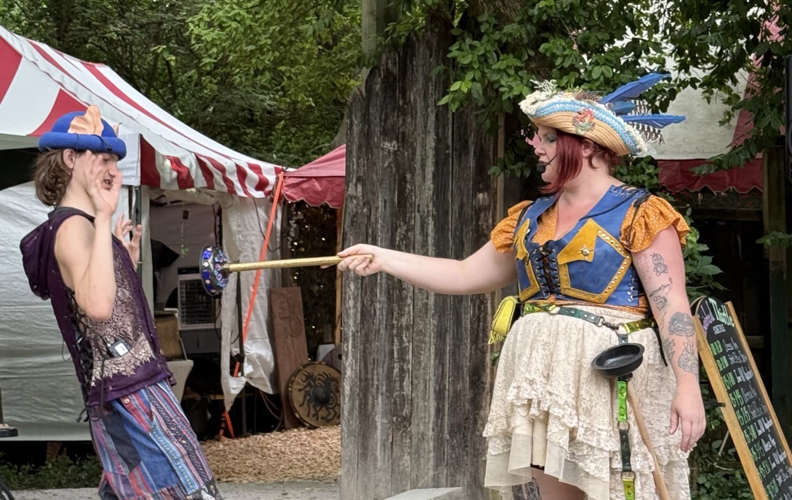 Captain Shh-nel in a feathered pirate hat and striped stockings levels a glitter-handled plunger at Stu of the Loo, who recoils, at an outdoor Renaissance faire