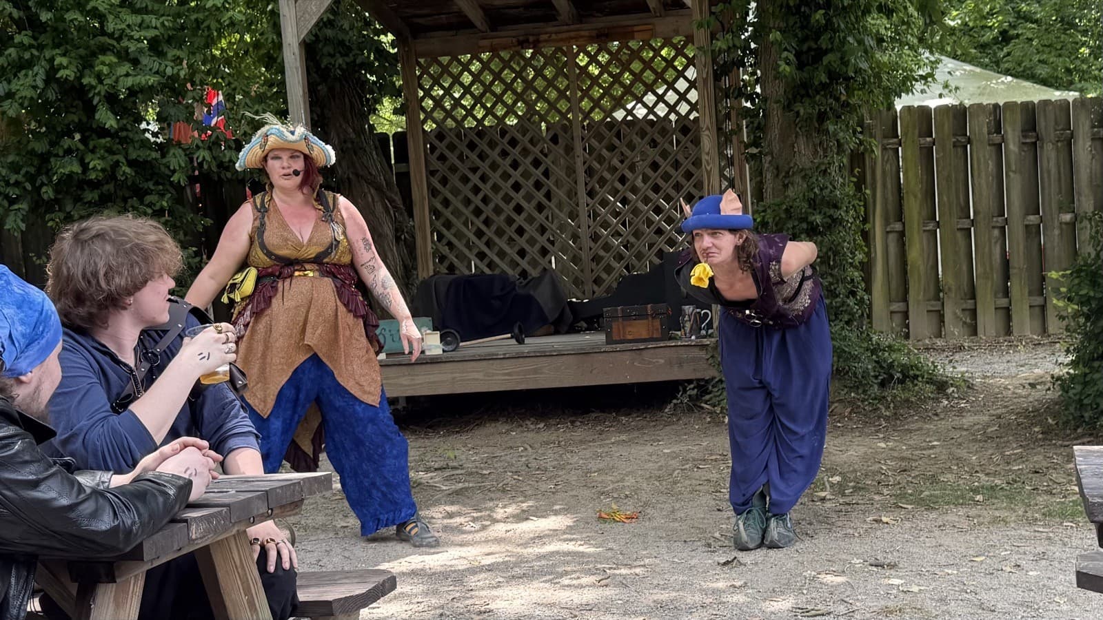Captain Shh-nel and Stu of the Loo mid-bit as Stu offers a yellow flower, with patrons watching from a picnic table at an outdoor faire