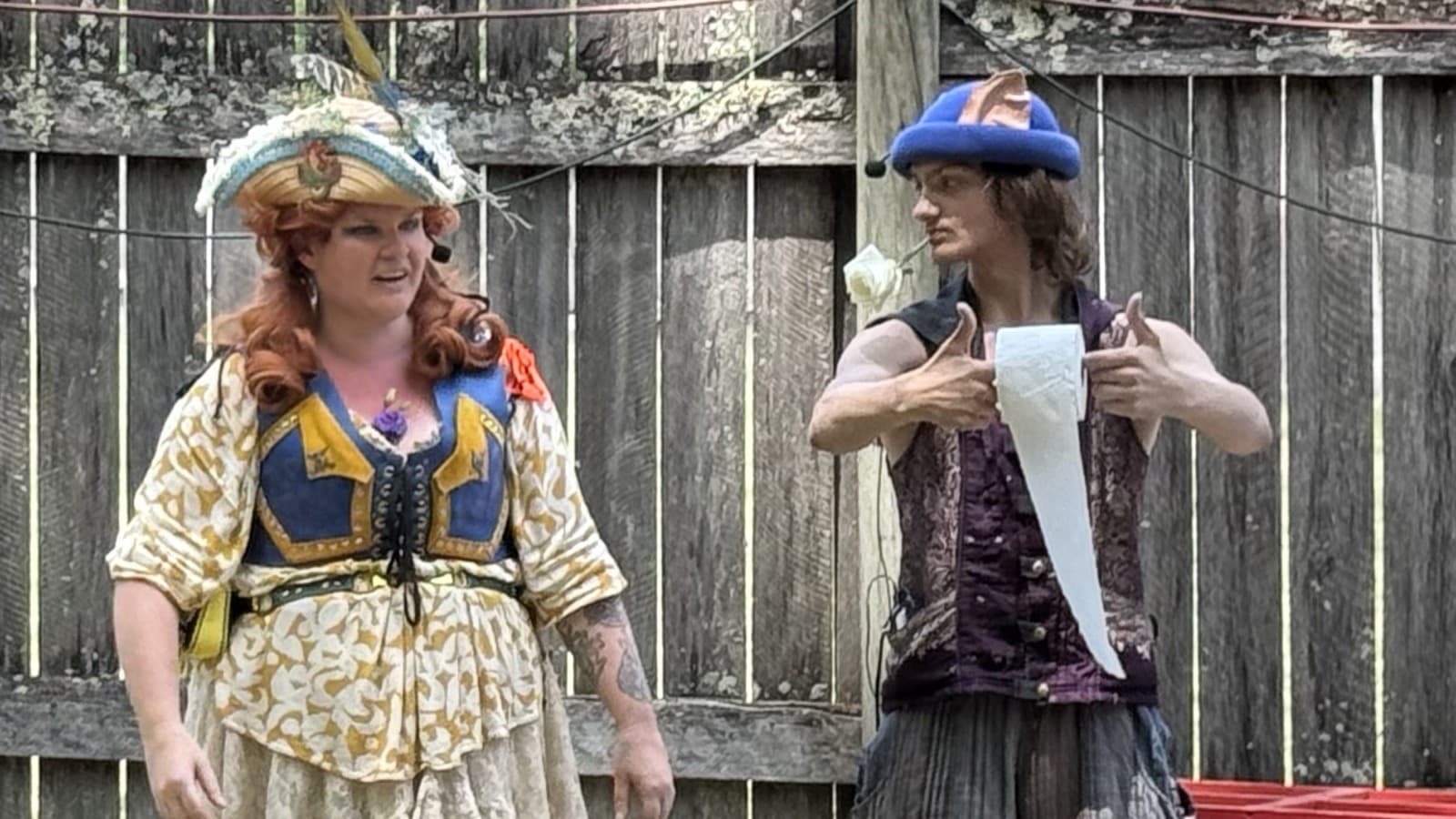 Captain Shh-nel and Stu of the Loo performing the Human Toilet Paper Holder bit, Stu holding out a roll of toilet paper, at an outdoor Renaissance faire