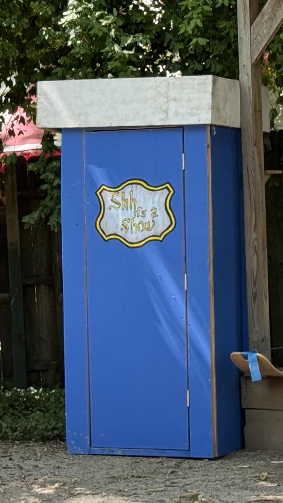 Old Blue, the show's tall blue theatrical outhouse, its door painted with a Shh It's A Show crest, standing outdoors among the trees