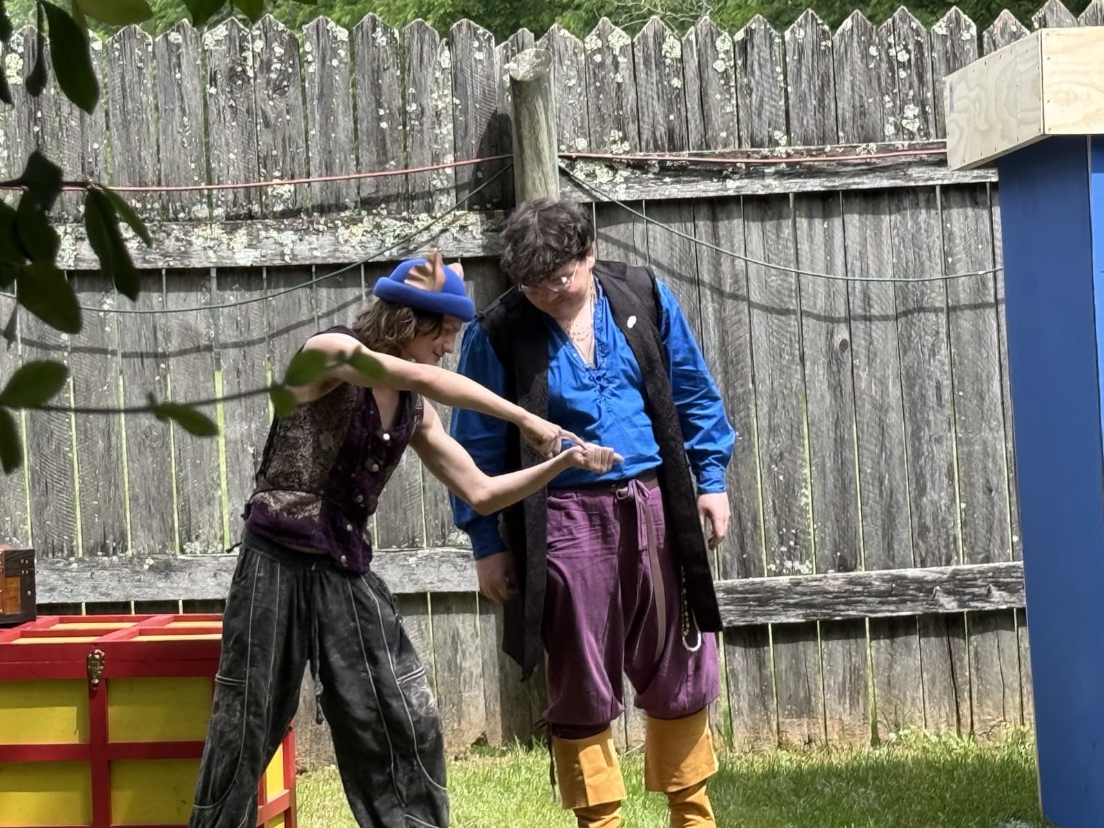Stu of the Loo works a sleight-of-hand trick on a seated volunteer's hand, a red and yellow prop chest beside them against a weathered fence