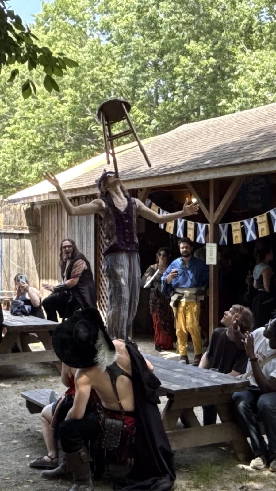 Stu of the Loo balances a wooden stool on his chin, arms outstretched, as a seated faire audience watches